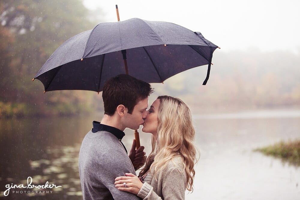 A couple hide under an umbrella and kiss during their rainy engagement session on the charles river in boston massachusetts