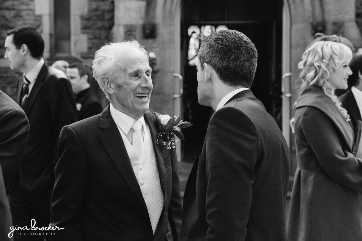 The father of the groom laughs with a friend outside the church of this classic vintage wedding