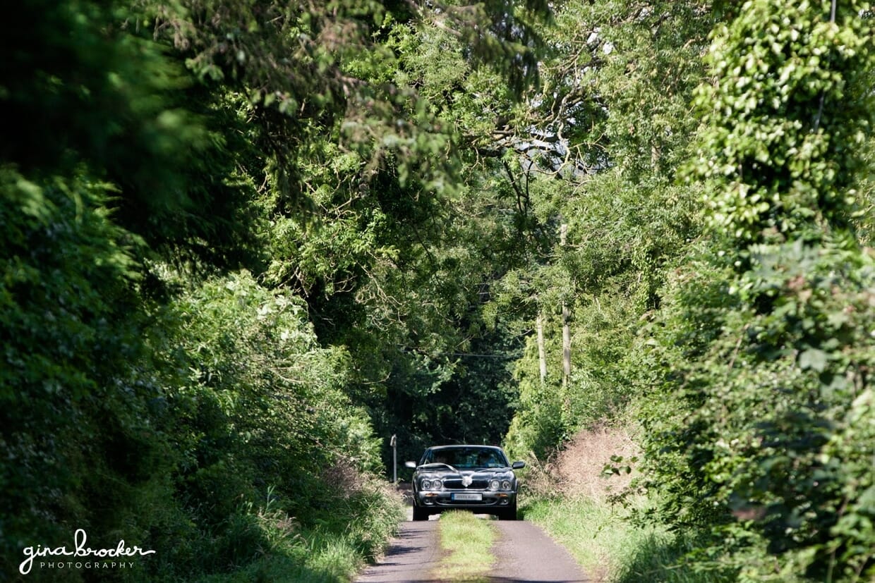 A vintage wedding car drives through a tree lined road on the way to an elegant wedding ceremony