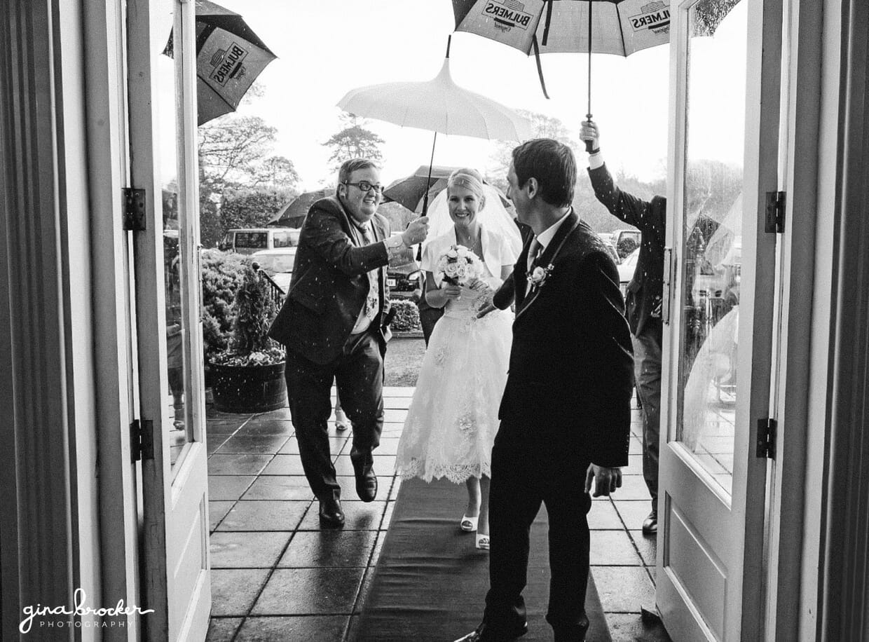 The bride and groom run into their venue with umbrellas as it starts to rain during their retro wedding