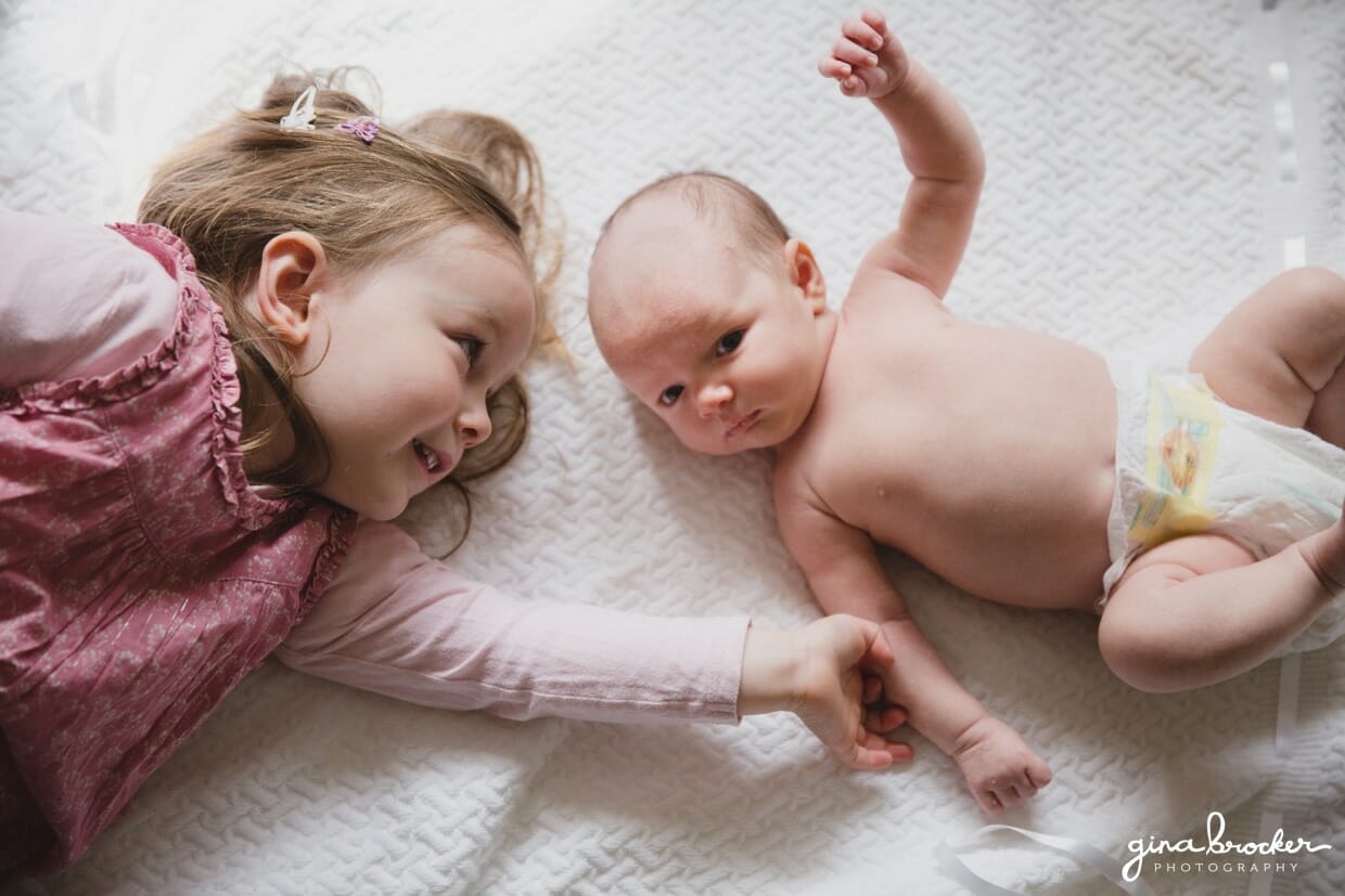 Toddler laying on a blanket and holding her baby sisters hand at family photo session at home in Boston