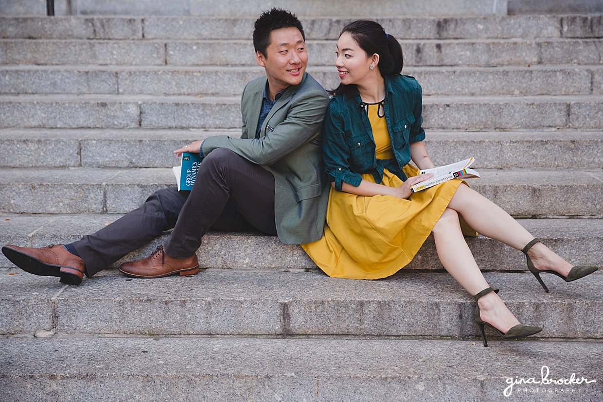 A couple sit back to back with books on the steps of Northeastern University during their University themed engagement in Boston, Massachusetts