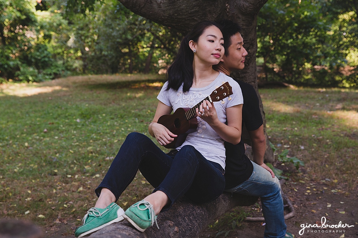 A couple sit together on a tree and play music during their Boston engagement session in Back Bay Fens