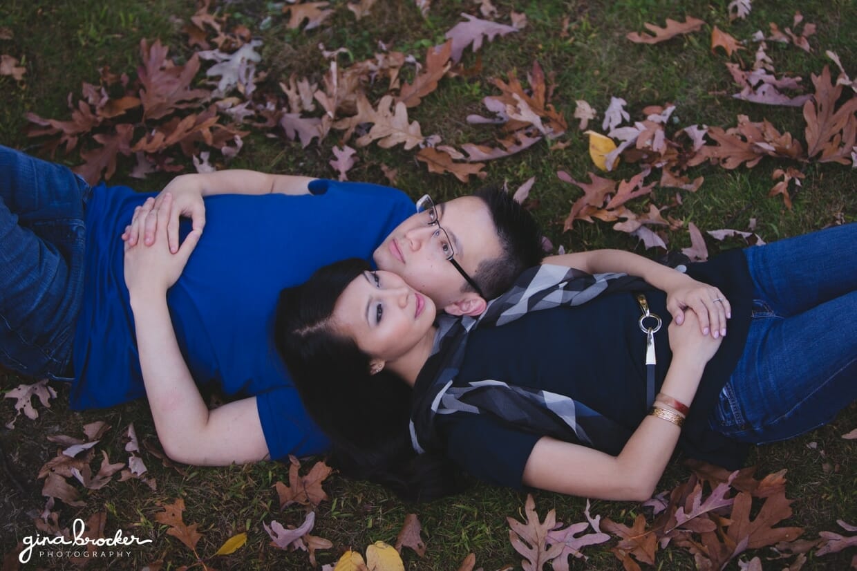 A couple lay together on leaves and look into the sky during their fall engagement session in boston
