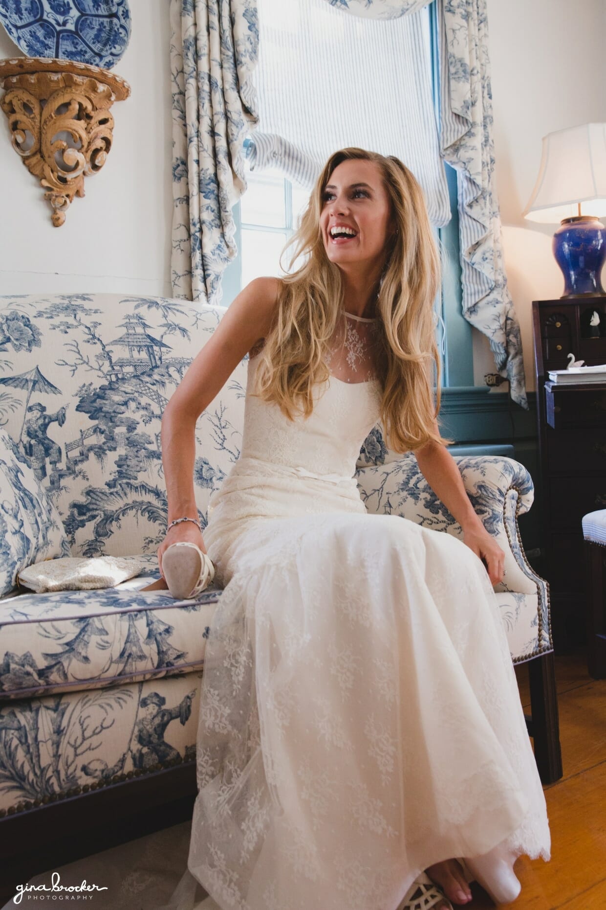 The bride laughs while she sits on the couch to put on her chanel wedding shoes before her Nantucket wedding at the Westmoor Club