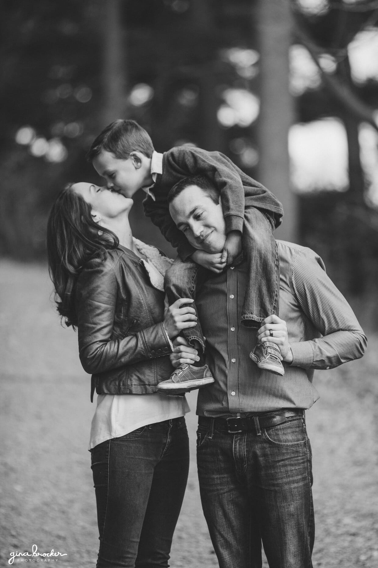 A sweet and funny photograph of a mother kissing her son while he rides on his father back during their family photo session in Arnold Arboretum, Boston