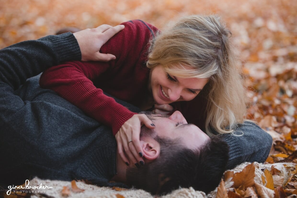 A super sweet photograph of a couple smiling at the each other as they lay on blanket in the fallen leaves during their fall couple session at the Arnold Arboretum in Boston, Massachusetts