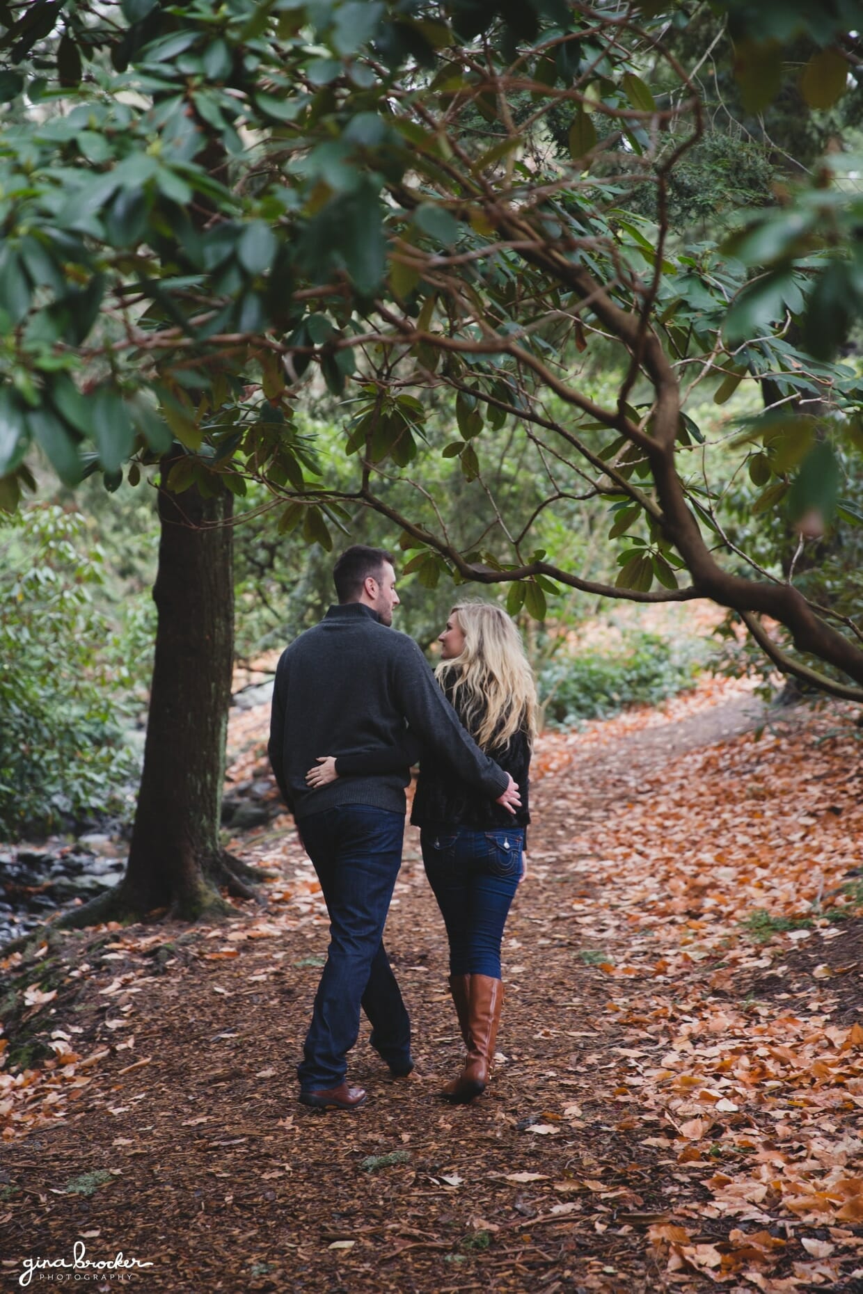 A photograph of a couple through a forest pathway with their arms around each other during their fall couple session in Boston's Arnold Arboretum