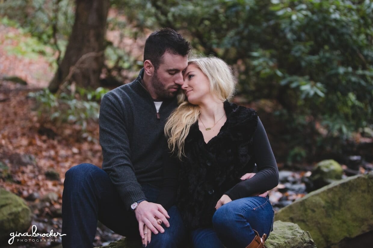 A sweet photograph of a couple sitting together on a rock in the woods during their fall couple session at the Arnold Arboretum in Boston, Massachusetts