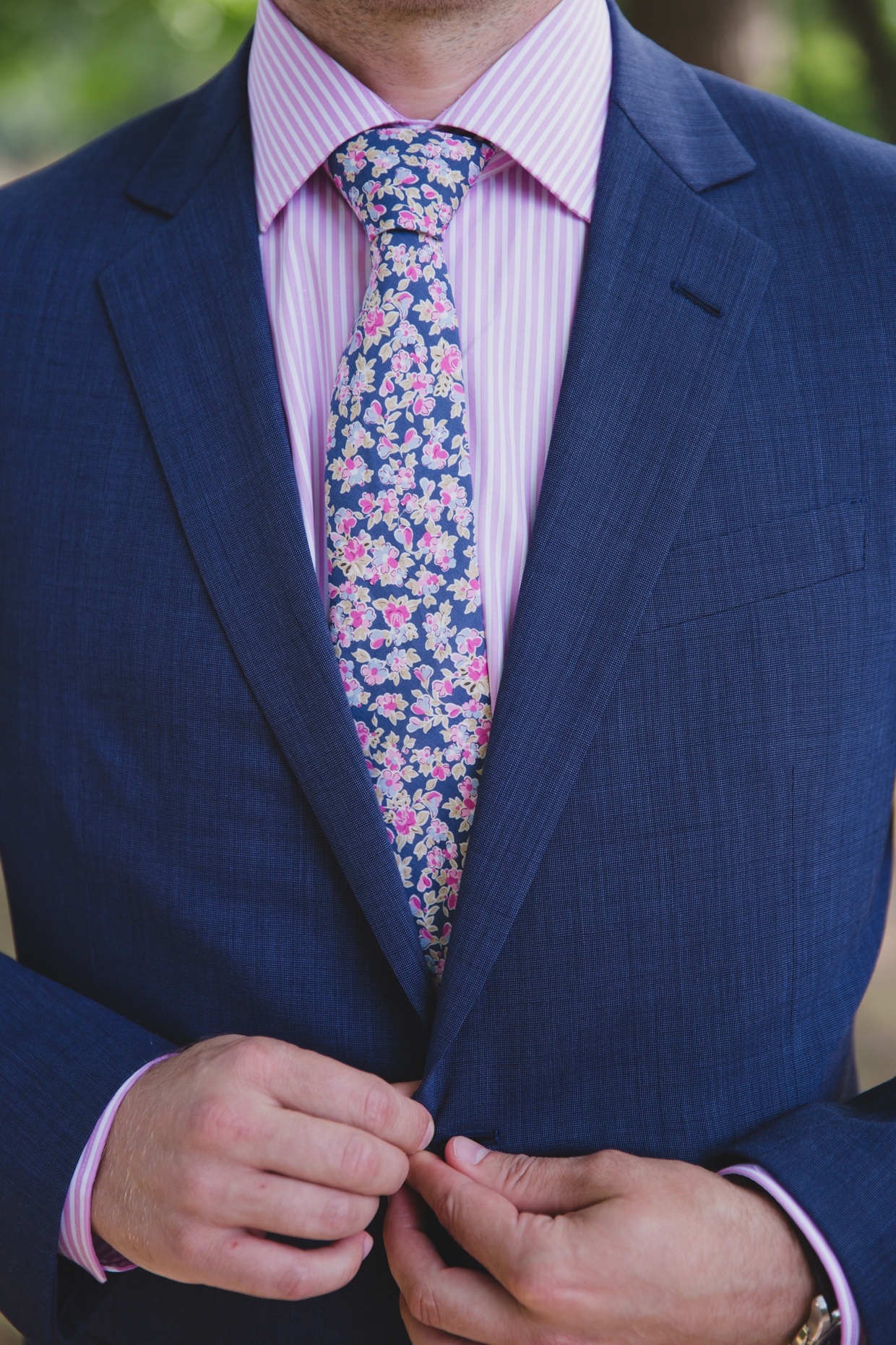 A detailed photograph of a groom buttoning his blue jacket with floral tie and pink pinstripe shirt before his backyard wedding