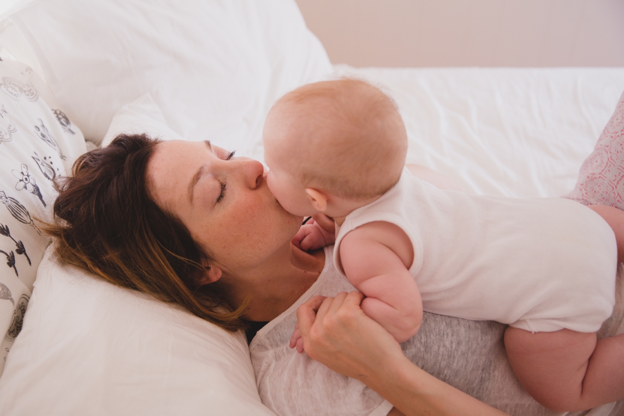 A sweet photograph of a mother kissing her baby as they lay in bed during an in home family photo session in Boston