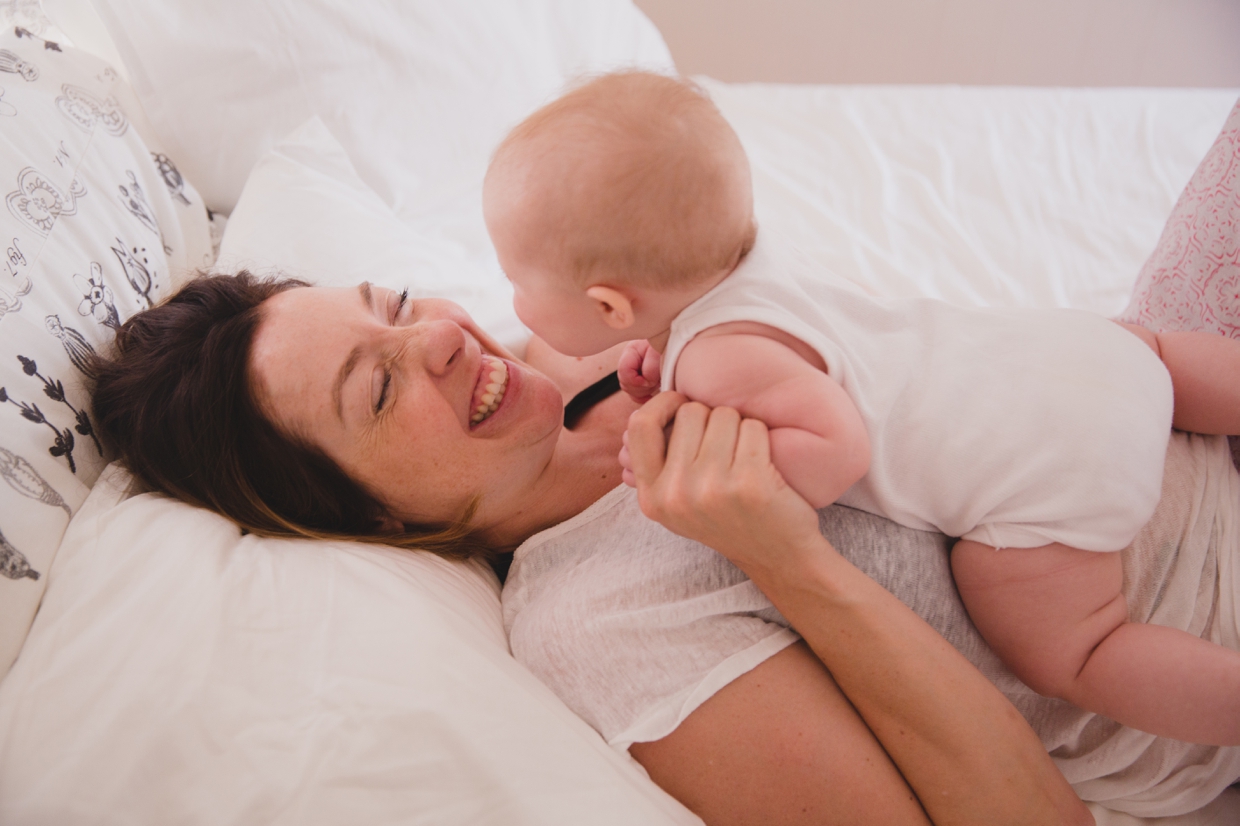 A cute photograph of mother laughing as she lays in bed with her baby during an in home family photo session in Boston
