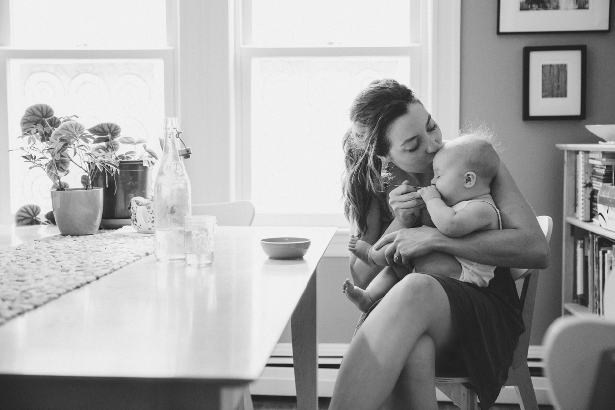 An intimate photograph of a mother kissing her baby while she feeds her at the table during an in home family photo session in Boston