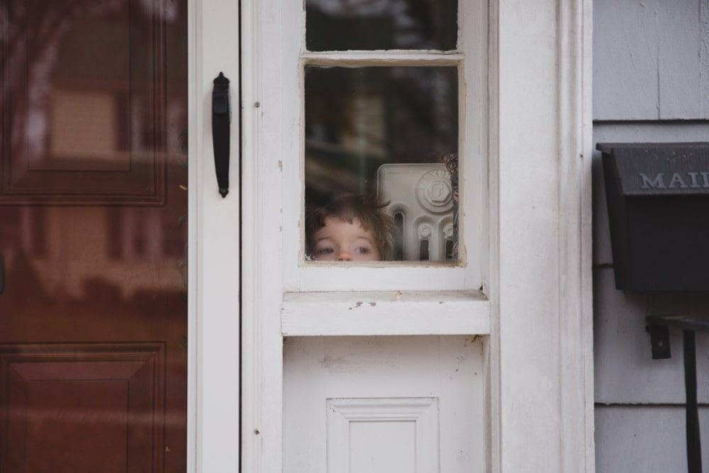 A documentary photograph of a little boy peeking out the window of his house during a in home lifestyle family session in Boston, Massachusetts