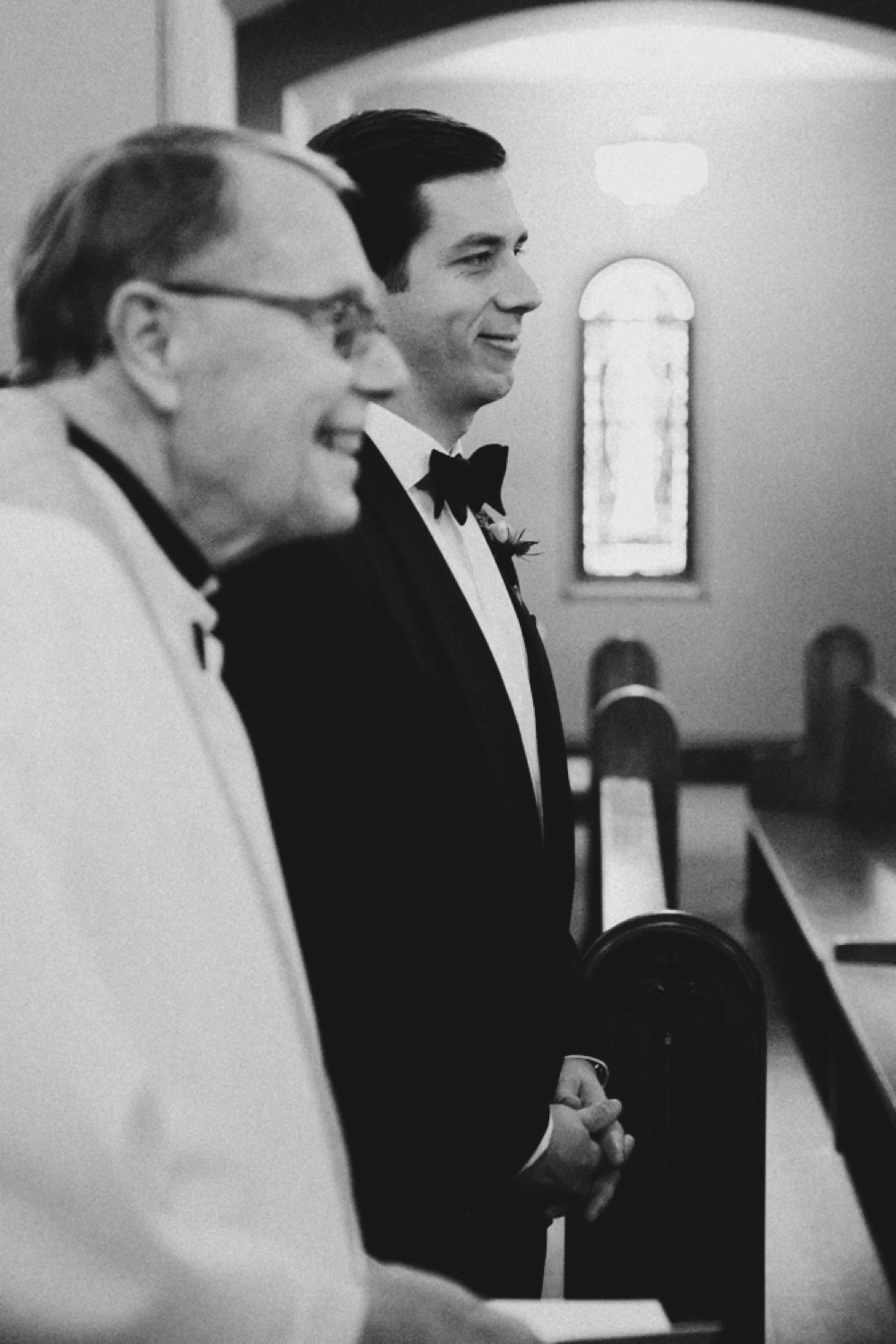 A photojournalistic portrait of a groom smiling as he watches his bride walk down the aisle of the St. Augustin's Church during a Newport, Rhode Island Wedding