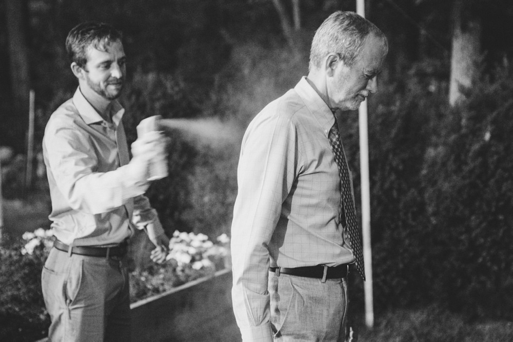 A documentary photograph of a wedding guest spraying his father with mosquito repellant at a backyard wedding in Boston, Massachusetts
