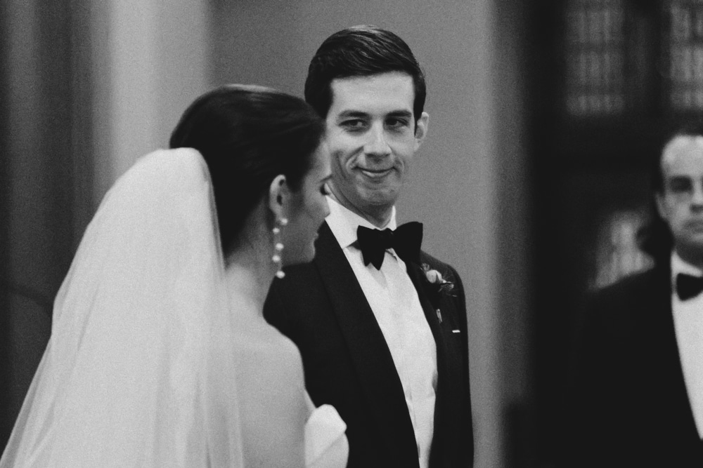 A documentary photograph of a groom smiling at his bride during their wedding ceremony at St. Augustin's Church in Newport, Rhode Island
