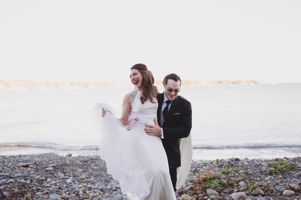 A documentary photograph of a groom carrying his bride over some rocks at the Oceanview of Nahant during their Massachusetts wedding