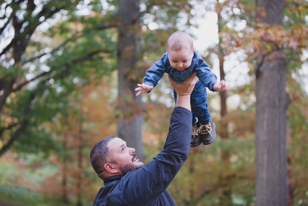 A lifestyle photograph of a father lifting his baby boy in the air during a fall family lifestyle mini session at the Arnold Arboretum in Boston, Massachusetts