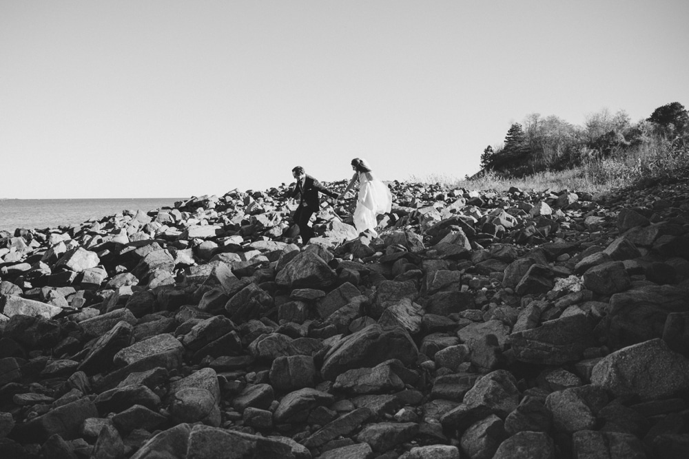 A documentary photograph of a couple walking on the rocks by the sea during their Massachusetts wedding at the Oceanview of Nahant