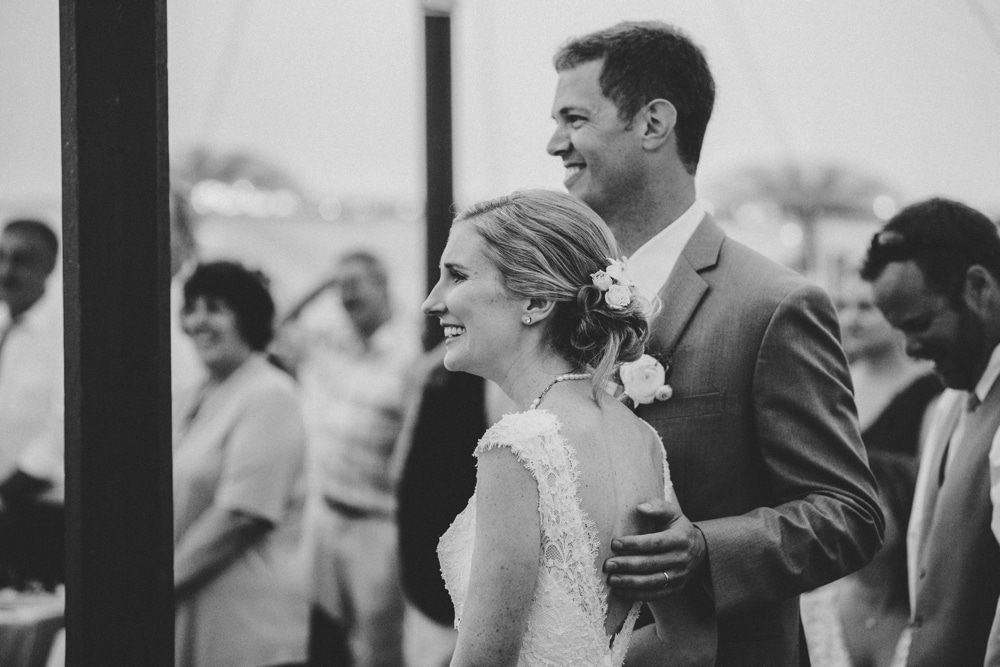 A documentary photograph of a bride and groom smiling during the wedding speeches at their fun summertime Cape Cod wedding at the Pilgrim's Monument in Provincetown, Massachusetts