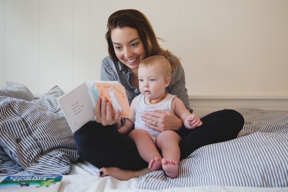 A intimate documentary photograph of a mother reading a book to her baby girl during a Boston in home lifestyle session