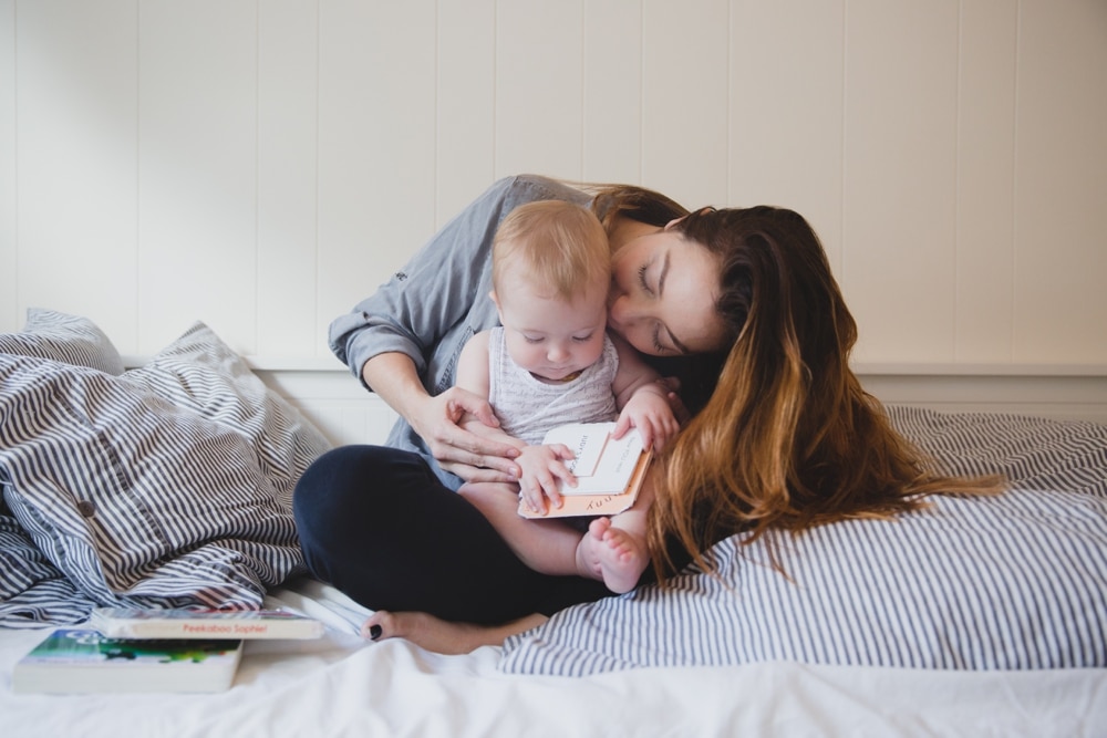 A photojournalist photograph of a mother kissing her baby girl while they read books in bed during their in home lifestyle session in Boston