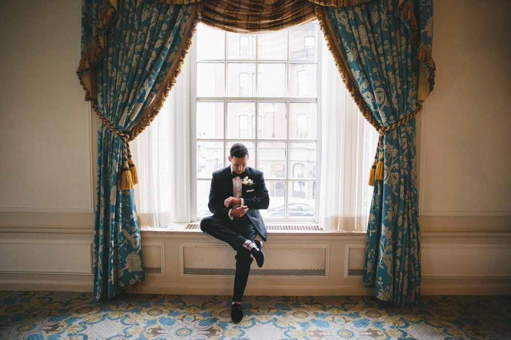 A documentary photograph of a Groom sitting by a window in the Taj Boston Hotel while he waits for his bride to arrive to their first look before their Boston City Wedding