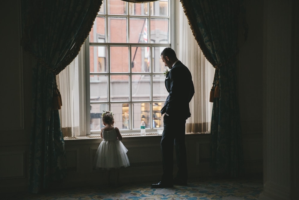 A documentary photograph of a flower girl and Groomsman looking out the window of the Taj Hotel during a Boston City Wedding