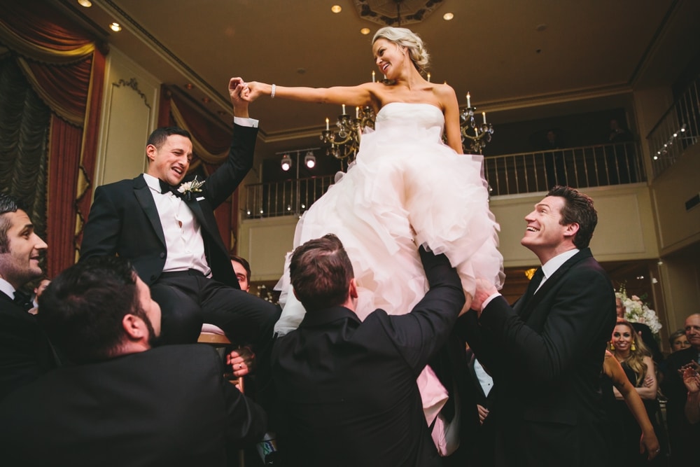 A documentary photograph of the bride and groom holding hands as they are lifted in chairs during the hora at their Taj Boston Hotel Wedding in Boston