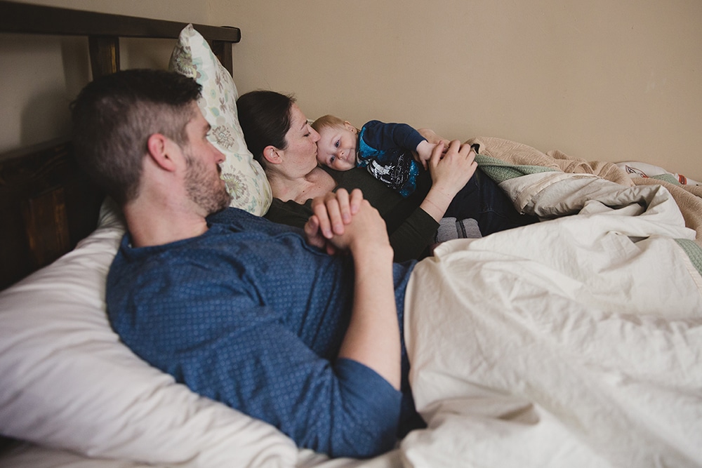 A documentary photograph of a father looking over at his wife kissing their son during an at home family session in Boston, Massachusetts