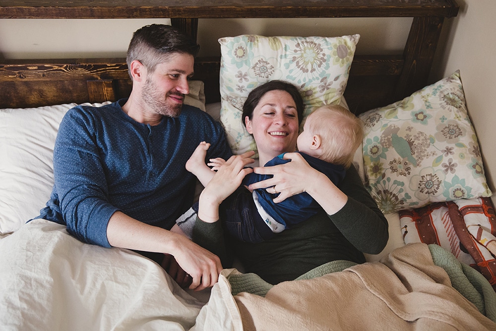 A documentary photograph of a father smiling at his wife and son as they snuggle in bed during a family session at home in Boston, Massachusetts