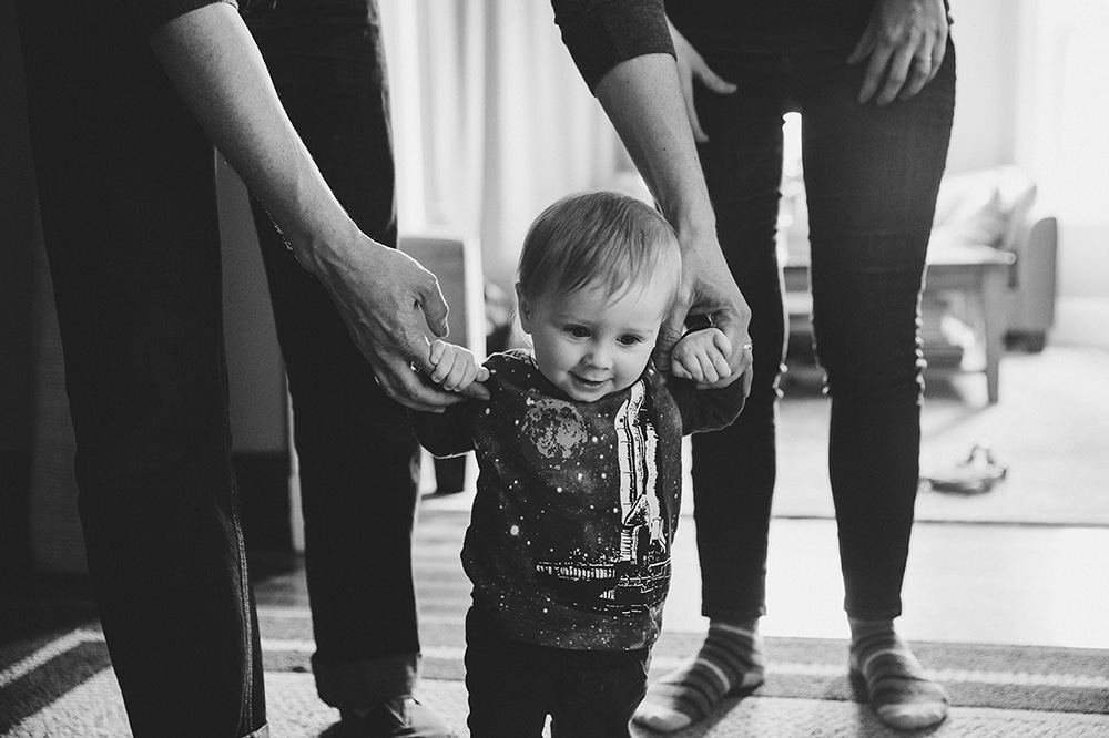 A documentary photograph of a father helping his baby boy walk during a family session at home in Boston, Massachusetts