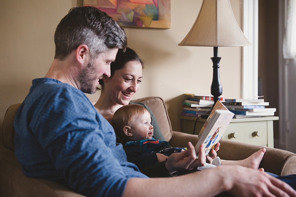 A documentary photograph of a mother and father reading a book to their baby boy during a family session at home in Boston, Massachusetts