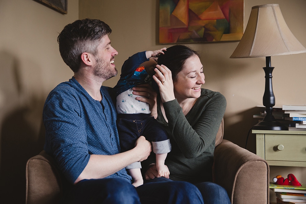 A documentary photograph of a baby boy grabbing his mother's hair as she laughs with her husband during a family session at home in Boston, Massachusetts