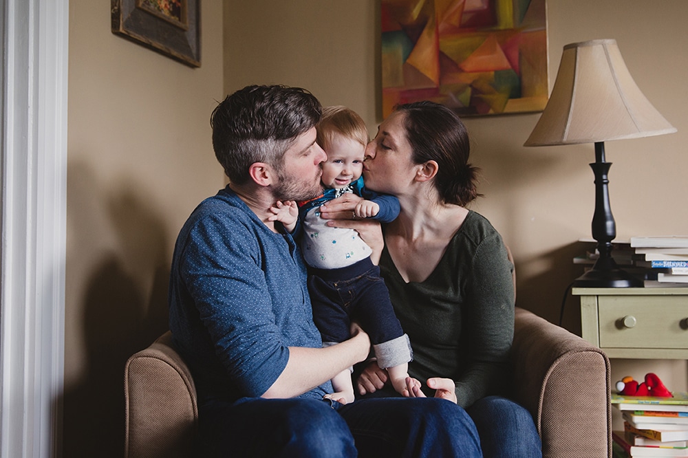 A documentary photograph of a mother and father kissing their baby boy during a family session at home in Boston, Massachusetts