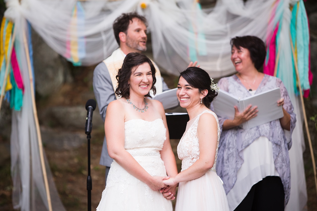 A documentary photograph of two brides looking out at their guests during their outdoor wedding ceremony at Friendly Crossways
