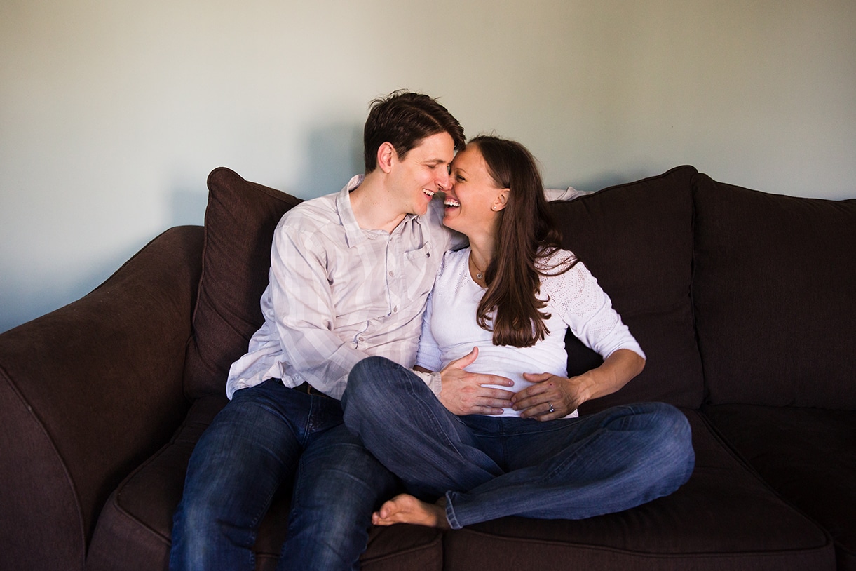 A lifestyle photograph of an expecting couple laughing together on the couch during their in home family session in Boston