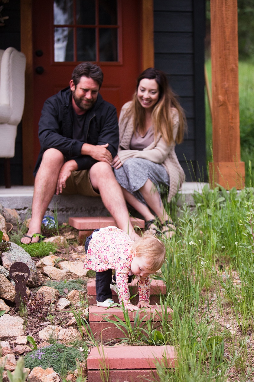 A lifestyle photograph of parents watching their daughter climb the steps to their porch during their evergreen family session at home in Colorado