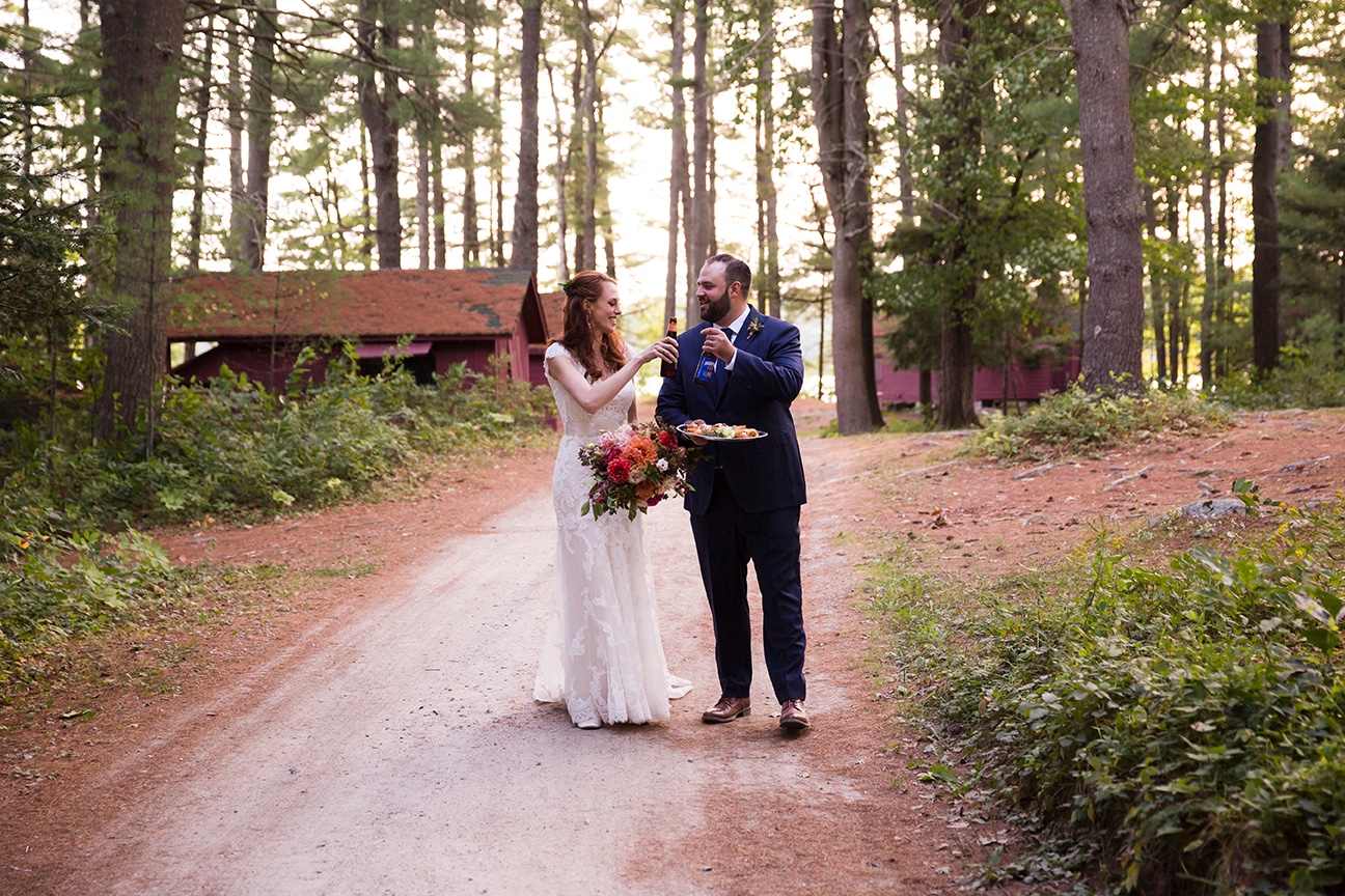 A documentary photograph of a bride and groom walking through the woods with craft beer during their Kingsley Pines Camp wedding in Maine