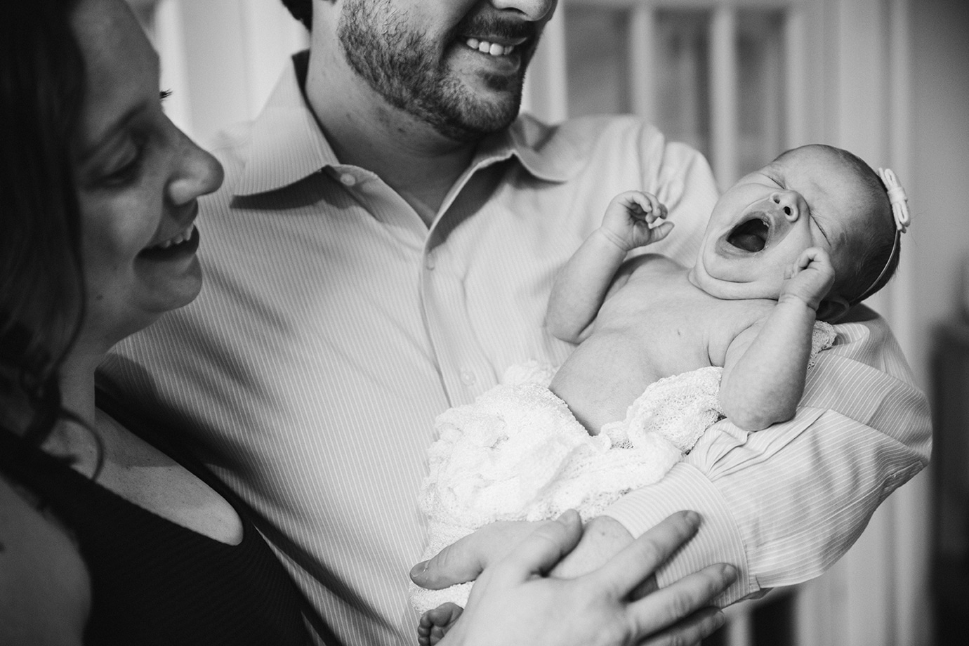 A lifestyle photograph of a mother and father holding their yawning baby girl during their in home newborn session in the Jamaica Plain neighbourhood of Boston