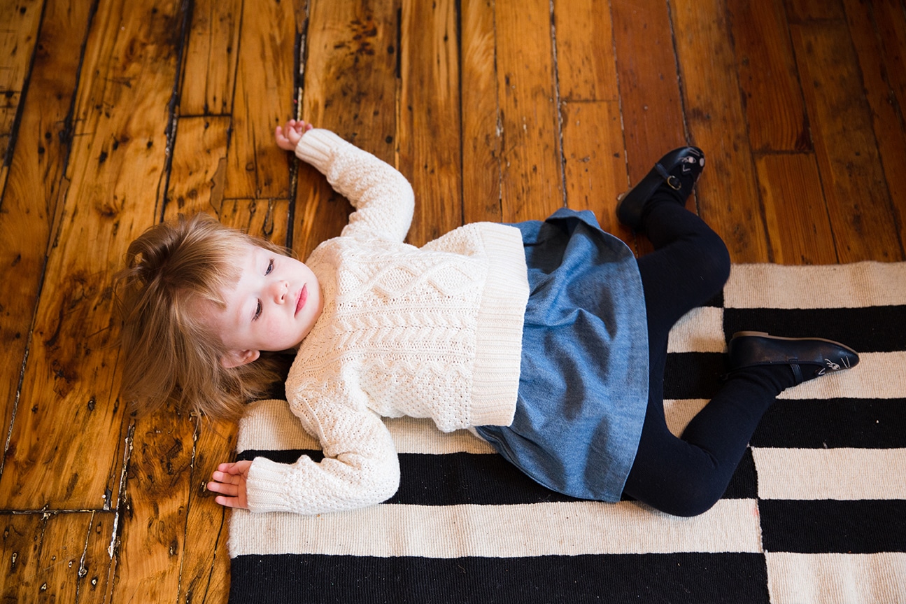 A lifestyle photograph of a toddler laying on the ground during an in home newborn session in the Jamaica Plain neighbourhood of Boston, Massachusetts