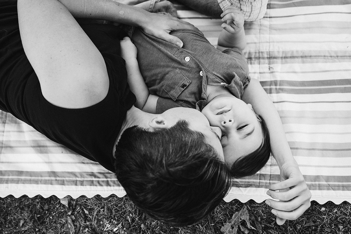A lifestyle portrait of a mother kissing her son as they lay on blanket during their Jamaica Pond family session