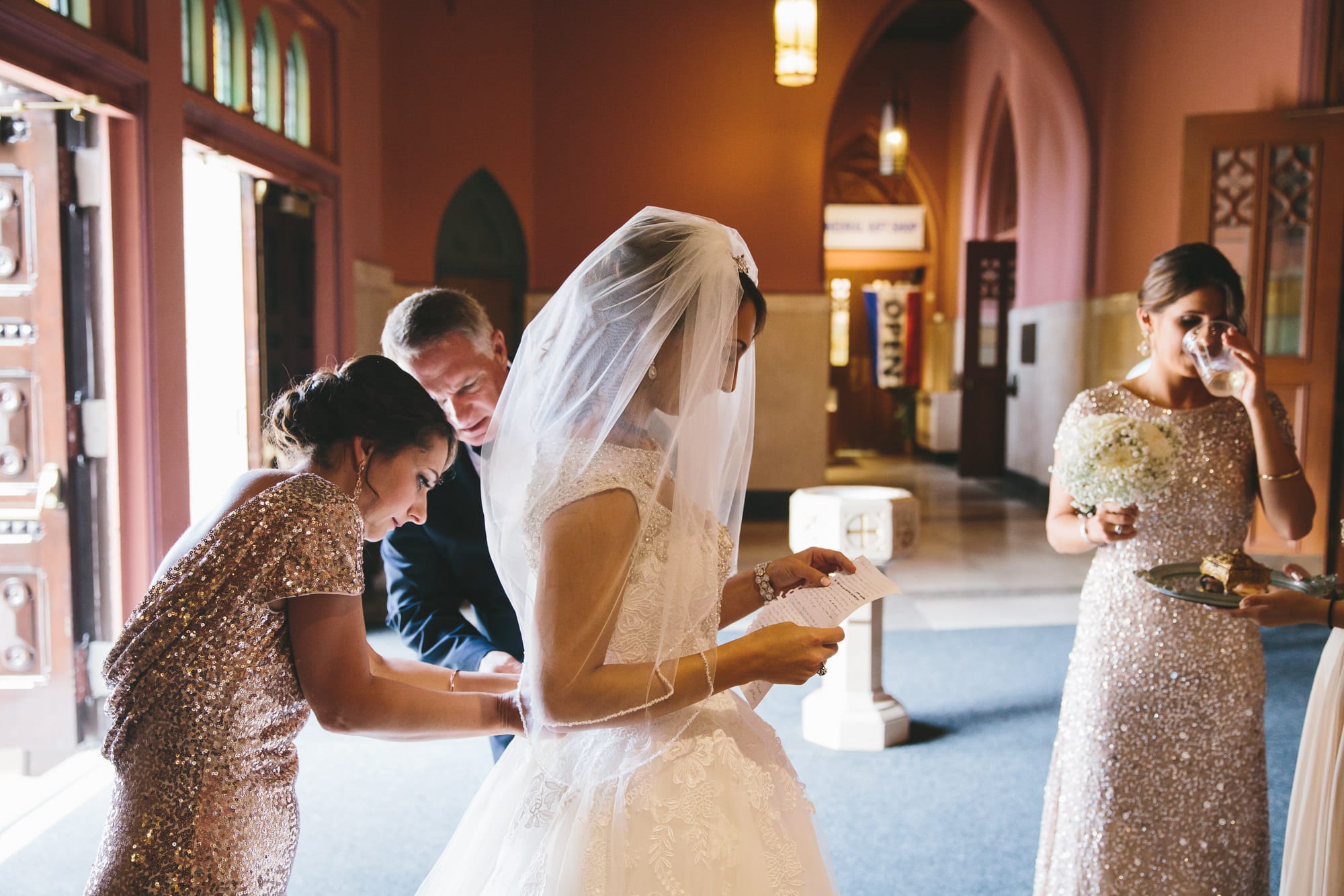 A documentary photograph of the brides father and sister fixing her dress while she reads a letter from her groom moments before walking down the aisle of the Cathedral of the Holy Cross during her State Room Wedding in Boston, Massachusetts