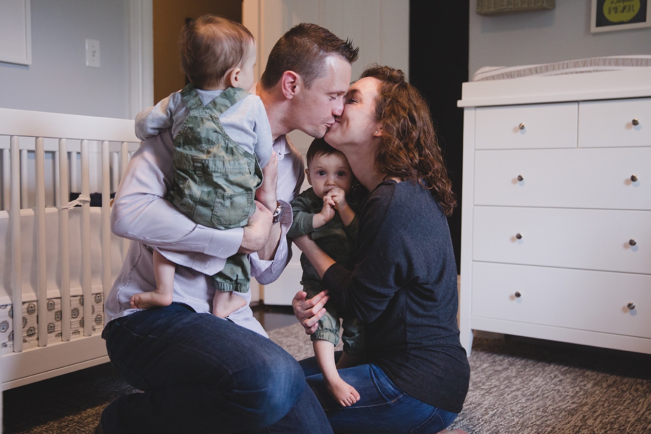A documentary photograph of parents kissing while holding their twin boys during an in home family session in Boston, Massachusetts
