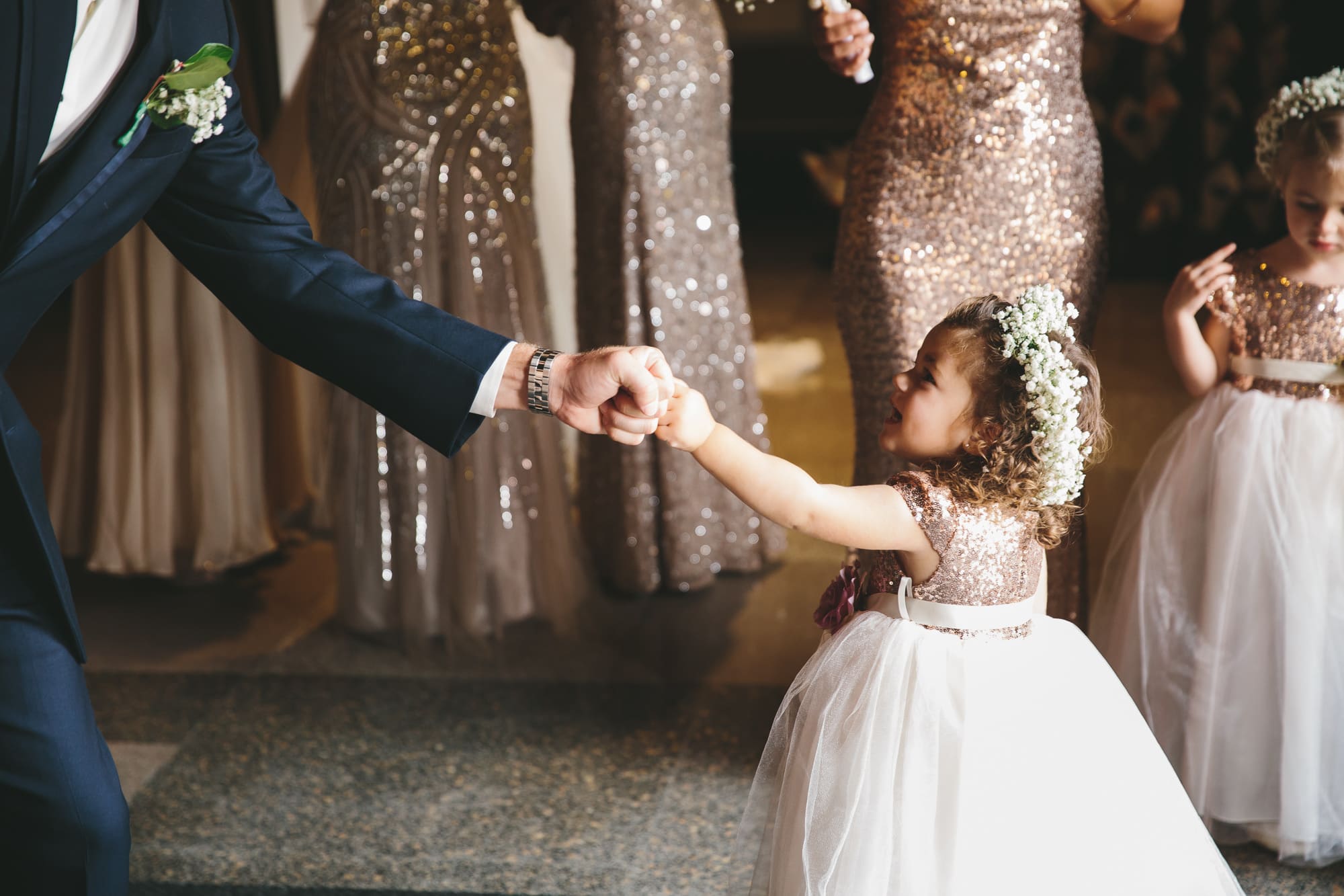 A documentary photograph of a flower girl fist bumping a groomsmen before they walk down the aisle of the Cathedral of the Holy Cross during a State Room Wedding