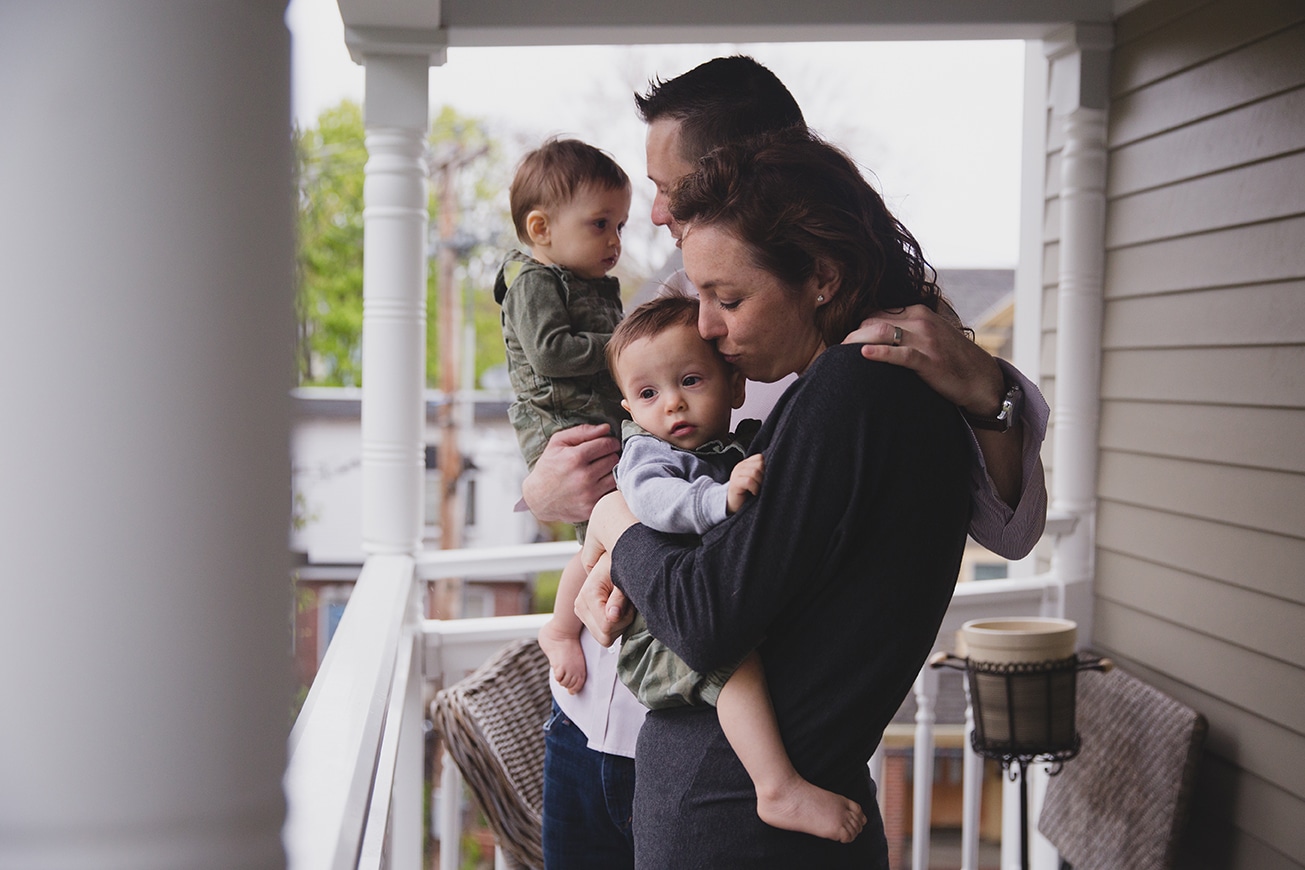 A documentary photograph of a mom kissing her baby boy as they stand on their porch during an in home family session in Boston, Massachusetts