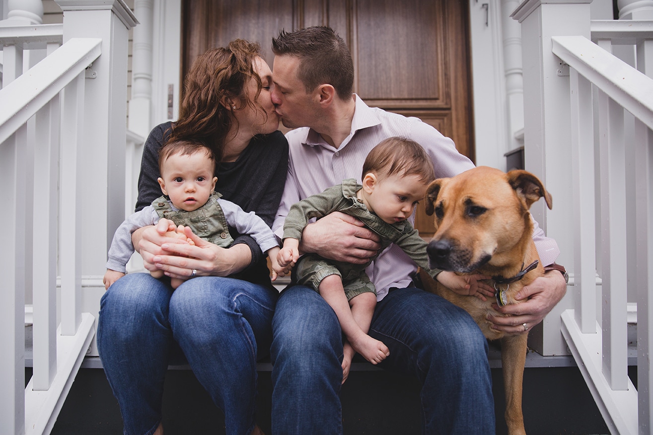 A documentary photograph of a parents kissing as they sit on the porch with their twin boys and dog during an in home family session in Boston, Massachusetts