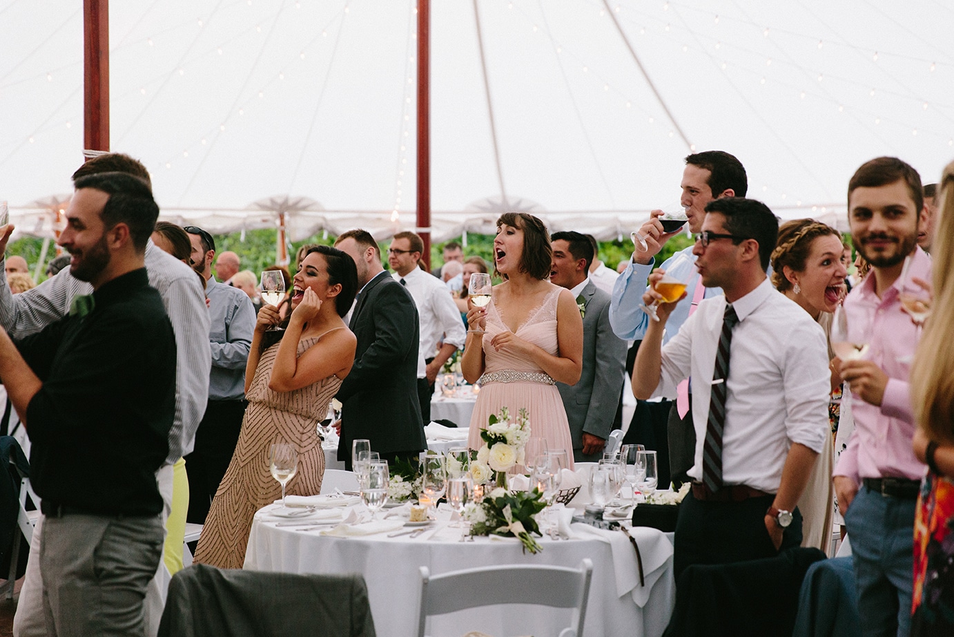 A documentary photograph of guests cheering the bride and groom as they enter the reception tent of their Moraine Farm Wedding in Beverly, Massachusetts