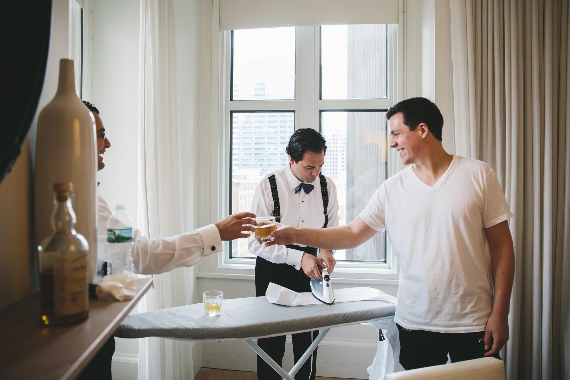 A groomsman hands the groom a drink while he gets ready for his State Room Wedding in the Ames Hotel
